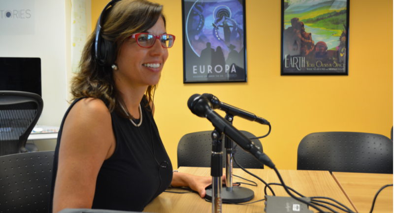 Rebecca Hester sits behind a desk with a microphone andconducts an interview in an episode of “In Place: Conversations About Displacement in the Commonwealth.”
